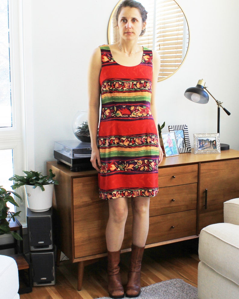 May include: A woman wearing a sleeveless red, green, yellow, and orange patterned dress with a scoop neck. She is standing in front of a wooden dresser with two drawers.