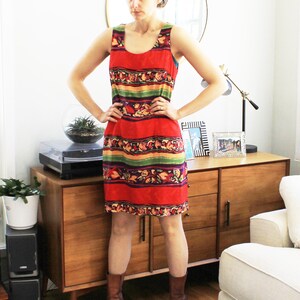 May include: A woman wearing a sleeveless red, green, and yellow patterned dress with brown boots. The dress has a horizontal stripe pattern with floral accents. The woman is standing in front of a wooden cabinet with a white couch in the background.