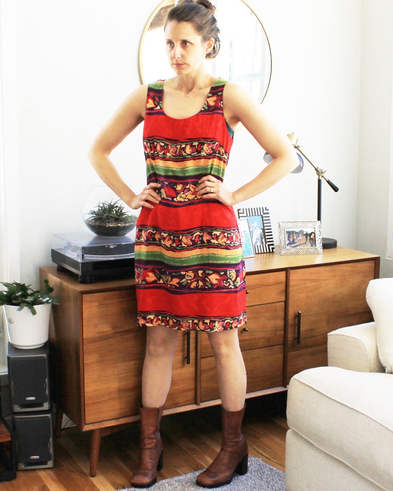 May include: A woman wearing a sleeveless red, green, and yellow patterned dress with brown boots. The dress has a horizontal stripe pattern with floral accents. The woman is standing in front of a wooden cabinet with a white couch in the background.