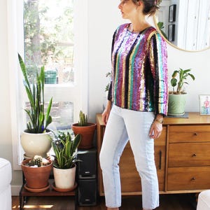 May include: A long-sleeved top covered in vertical stripes of multicoloured sequins in shades of purple, gold, teal, and black. The top is paired with light-wash denim jeans and brown leather slip-on shoes, creating a complete outfit ensemble.