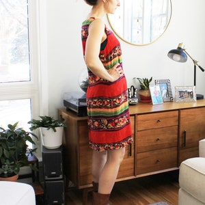May include: A woman wearing a sleeveless red, green, yellow, and purple patterned dress with brown ankle boots. The dress has a geometric pattern and is knee-length. The woman is standing in front of a wooden dresser with three drawers.