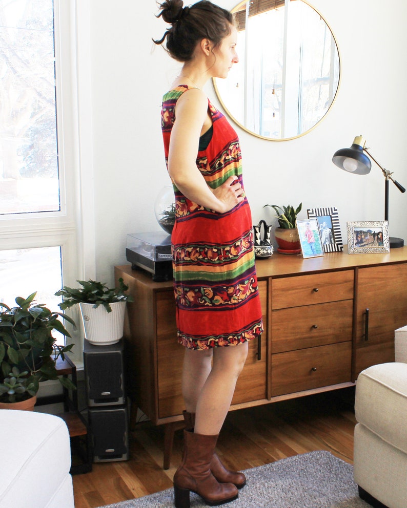 May include: A woman wearing a sleeveless red, green, yellow, and purple patterned dress with brown ankle boots. The dress has a geometric pattern and is knee-length. The woman is standing in front of a wooden dresser with three drawers.