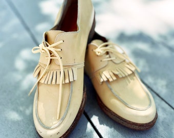 May include: A pair of light beige leather shoes with fringe detailing and laces. The shoes have a white sole and are sitting on a wooden surface.