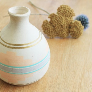 May include: A small, white ceramic vase with a round body and a narrow neck. The vase is decorated with gold and blue stripes and a peach-colored design.