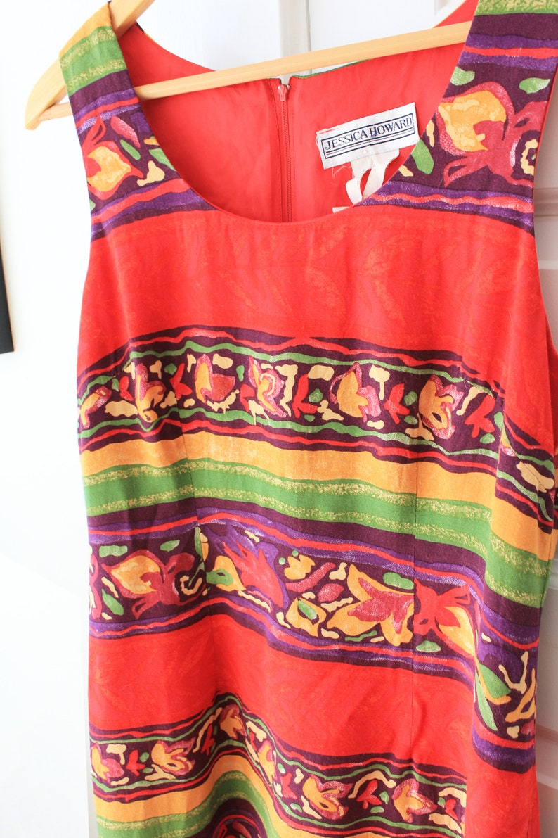 May include: A red sleeveless dress with a colorful floral and geometric pattern. The dress has a scoop neck and a zipper closure in the back. The dress has a label that reads "Jessica Howard!"