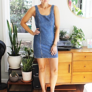 May include: A blue denim mini dress featuring a scoop neckline, thick shoulder straps, and vertical seam detailing running down the front. The dress has a fitted silhouette and ends above the knee, paired with two-toned clogs in pink and grey.