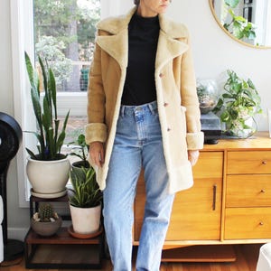 May include: A tan suede coat with a cream-colored faux shearling lining, wide lapels, and cuffs. The coat is worn over a black mock neck top and light blue denim jeans. The ensemble is completed with tan slip-on shoes with a buckle detail.