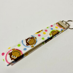 May include: A white fabric keychain with a colorful monkey print. The keychain has a silver metal ring.