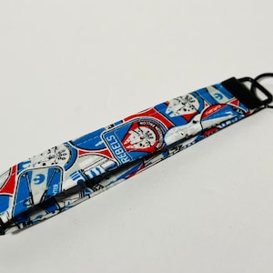 Pode incluir: Um chaveiro de tecido branco, azul e vermelho com tema Star Wars. O chaveiro apresenta imagens do Millennium Falcon, símbolos da Aliança Rebelde e o texto "REBEL 1".