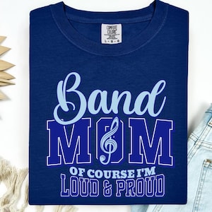 Puede incluir: Una camiseta azul marino con las palabras "Band Mom" en escritura azul claro y letras de bloque azules en negrita. El texto "Of course I'm Loud & Proud" está debajo. La camiseta tiene una etiqueta Comfort Colors.