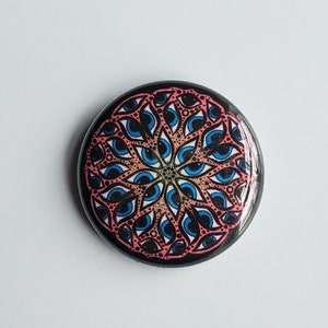 Könnte beinhalten: Ein runder Ansteckbutton mit einem sich wiederholenden radialen Muster aus blauen Augen mit schwarzen Pupillen und rosafarbenen Lidern. Das Design ist in einer symmetrischen, mandalaartigen Formation auf schwarzem Grund angeordnet.
