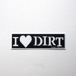 May include: A rectangular black sticker with white text that reads I heart DIRT, featuring a white heart symbol in place of the word love. The text Tender Caballero is printed in a smaller white font in the bottom right corner of the sticker.