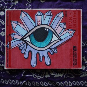 May include: A red USPS Priority Mail sticker with a hand-drawn illustration of an eye with blue and green colors. The eye is surrounded by pink and blue crystals. The text "PRIORI MAIL" and "FROM:" are visible on the sticker.
