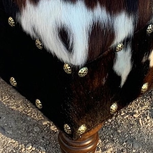 May include: A brown and white cowhide ottoman with gold-toned studs and wooden legs. The ottoman is sitting on a gravel surface.
