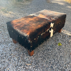 May include: A brown and white cowhide ottoman with decorative metal studs. The ottoman has a rectangular shape and is sitting on a gravel surface.