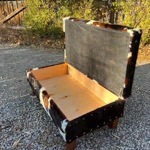 May include: A brown and white cowhide storage bench with a hinged lid. The lid is open, revealing the empty interior of the bench.