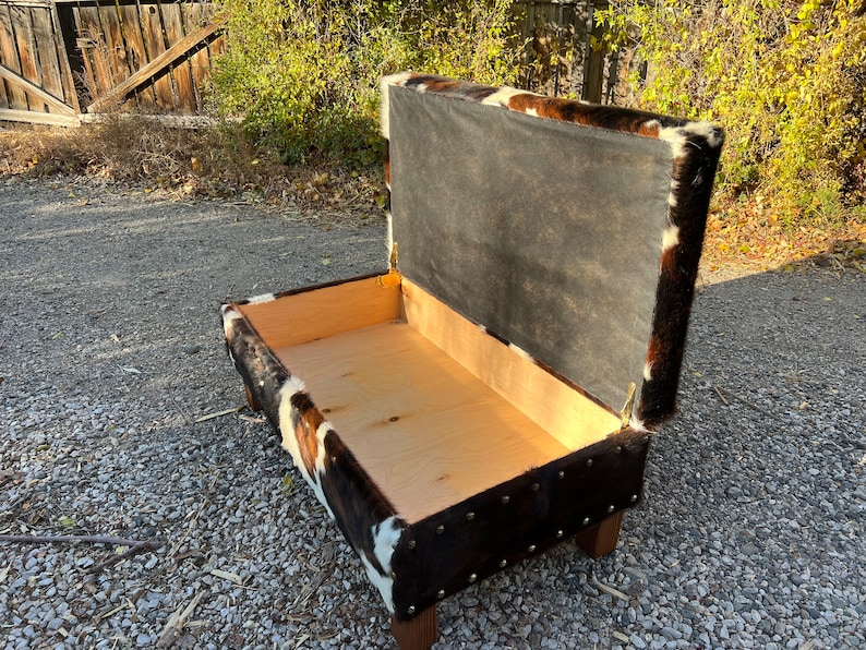 May include: A brown and white cowhide storage bench with a hinged lid. The lid is open, revealing the empty interior of the bench.