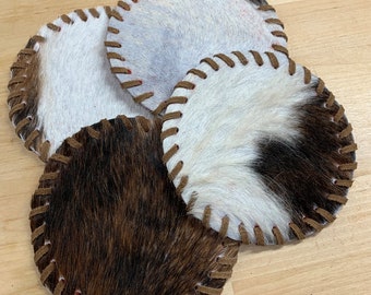 May include: Four brown and white cowhide coasters with brown leather stitching around the edges.