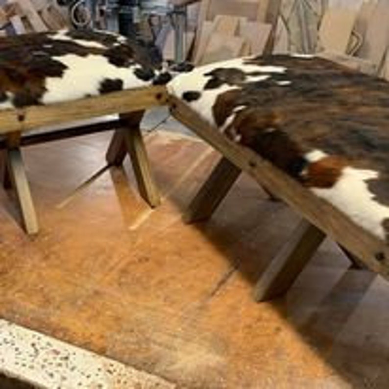 Pode incluir: Dois banquinhos de madeira com um assento de couro de vaca marrom e branco. Os banquinhos t&ecirc;m um design simples e r&uacute;stico com uma base em forma de cruz.