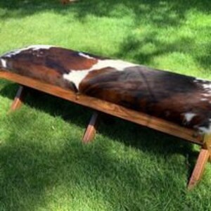 Pode incluir: Um banco de madeira r&uacute;stico com um assento de couro de vaca. O banco tem um acabamento de madeira natural e o couro de vaca &eacute; castanho e branco.