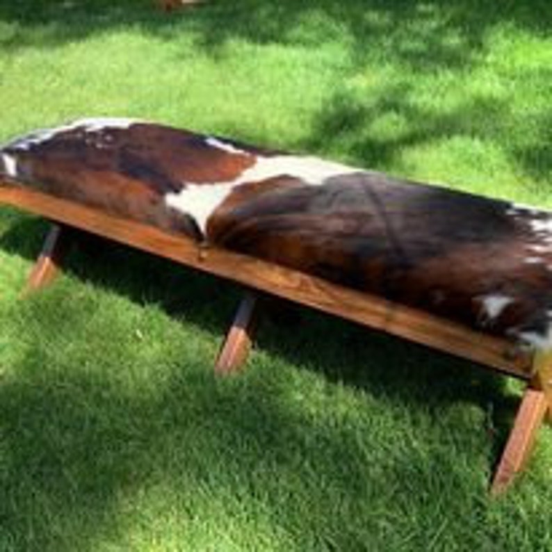 Pode incluir: Um banco de madeira r&uacute;stico com um assento de couro de vaca. O banco tem um acabamento de madeira natural e o couro de vaca &eacute; castanho e branco.