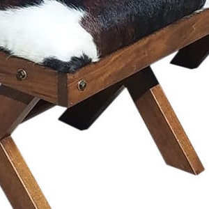 Pode incluir: Um banquinho de madeira com um assento de couro de vaca preto e branco. O banquinho tem um design simples e r&uacute;stico com quatro pernas que se cruzam em forma de X.