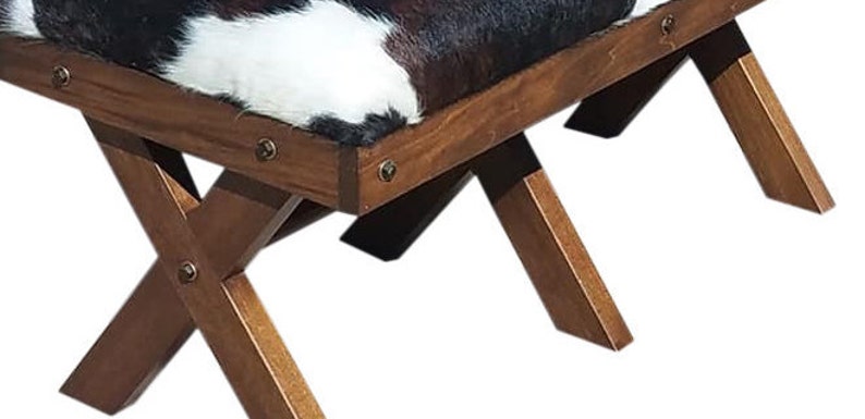 Pode incluir: Um banquinho de madeira com um assento de couro de vaca preto e branco. O banquinho tem um design simples e r&uacute;stico com quatro pernas que se cruzam em forma de X.