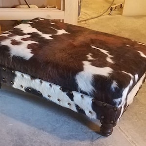 May include: A brown and white cowhide ottoman with a tufted top and studded trim. The ottoman has four wooden legs.