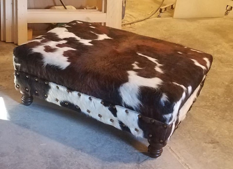 May include: A brown and white cowhide ottoman with a tufted top and studded trim. The ottoman has four wooden legs.