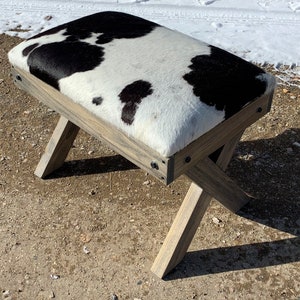 May include: A rustic wooden bench with a black and white cowhide seat. The bench has a simple, X-shaped base and is made of weathered wood.