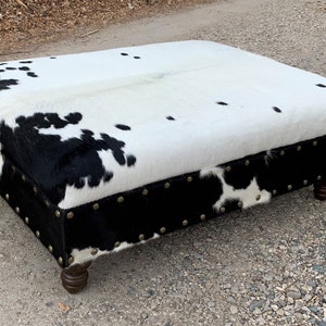 May include: A black and white cowhide ottoman with a black leather trim and brass studs. The ottoman has four wooden legs.
