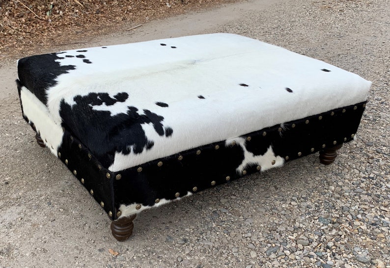 May include: A black and white cowhide ottoman with a black leather trim and brass studs. The ottoman has four wooden legs.