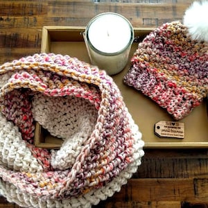 Peut inclure: Un ensemble de foulard et bonnet en crochet de couleur rose, blanc et orange. Le foulard est plié en deux et le bonnet est surmonté d'un pompon blanc. Un petit panneau en bois avec le texte "Merci de soutenir les petites entreprises" est en arrière-plan.