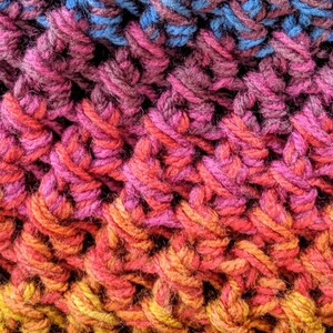 May include: Close-up of a crocheted fabric with a gradient of colors. The top section is blue and green, transitioning to shades of pink, orange, and yellow at the bottom. The texture is thick and textured, with visible stitches.