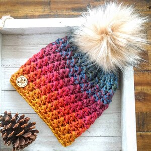 May include: A handmade crochet hat with a gradient of colors from yellow to blue, topped with a large, fluffy, light brown faux fur pom. A wooden button with a floral design is sewn on the side.