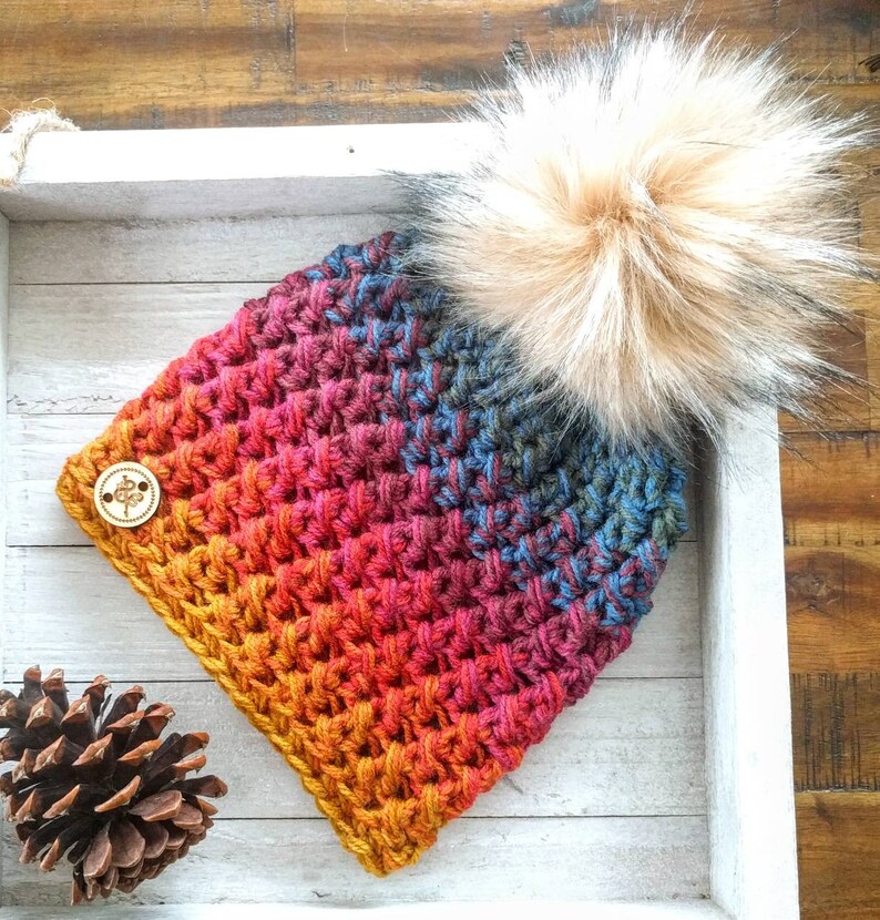 May include: A handmade crochet hat with a gradient of colors from yellow to blue, topped with a large, fluffy, light brown faux fur pom. A wooden button with a floral design is sewn on the side.