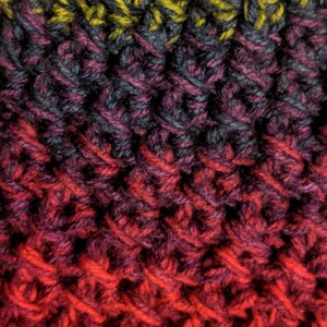 May include: Close-up of a knitted hat with a gradient of colours, starting with a dark grey and yellow, transitioning to purple, then red, and ending with orange and yellow. The hat has a fuzzy pom-pom on top.