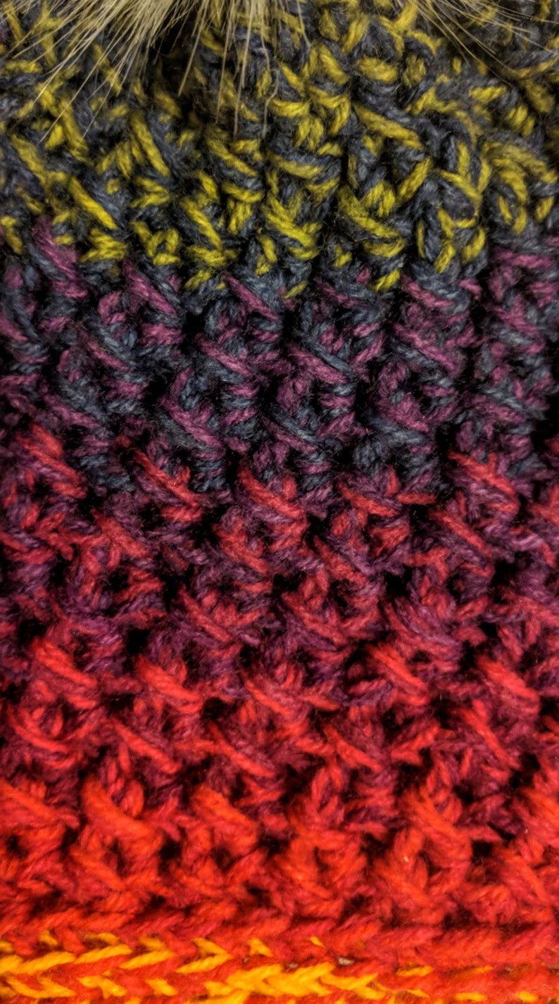 May include: Close-up of a knitted hat with a gradient of colours, starting with a dark grey and yellow, transitioning to purple, then red, and ending with orange and yellow. The hat has a fuzzy pom-pom on top.