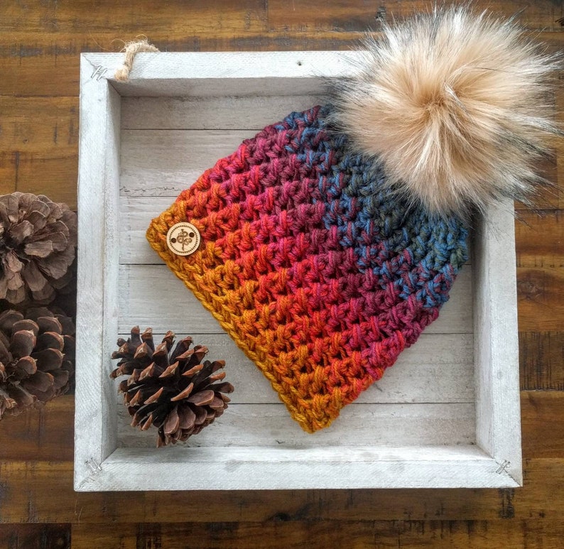 May include: A handmade, colorful crochet beanie with a faux fur pom-pom. The hat features a gradient of colors, including orange, red, blue, and purple. A small wooden button is attached to the side. The beanie is displayed in a white wooden tray.