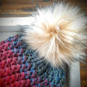 May include: A handmade crochet hat in shades of red, blue, and green, topped with a large, fluffy faux fur pom-pom in tan and gray. The hat's textured stitch creates a cozy, warm appearance. The background is a wooden surface.