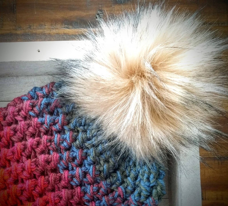 May include: A handmade crochet hat in shades of red, blue, and green, topped with a large, fluffy faux fur pom-pom in tan and gray. The hat's textured stitch creates a cozy, warm appearance. The background is a wooden surface.