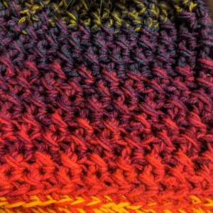 May include: Close-up of a crocheted hat with a gradient of colours, starting with dark purple and transitioning to red and yellow.