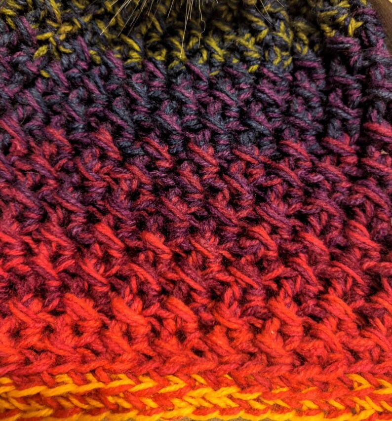 May include: Close-up of a crocheted hat with a gradient of colours, starting with dark purple and transitioning to red and yellow.