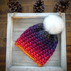 May include: A crocheted beanie with a gradient of red, orange, yellow, purple, and blue colours. The beanie has a white faux fur pom pom on top.