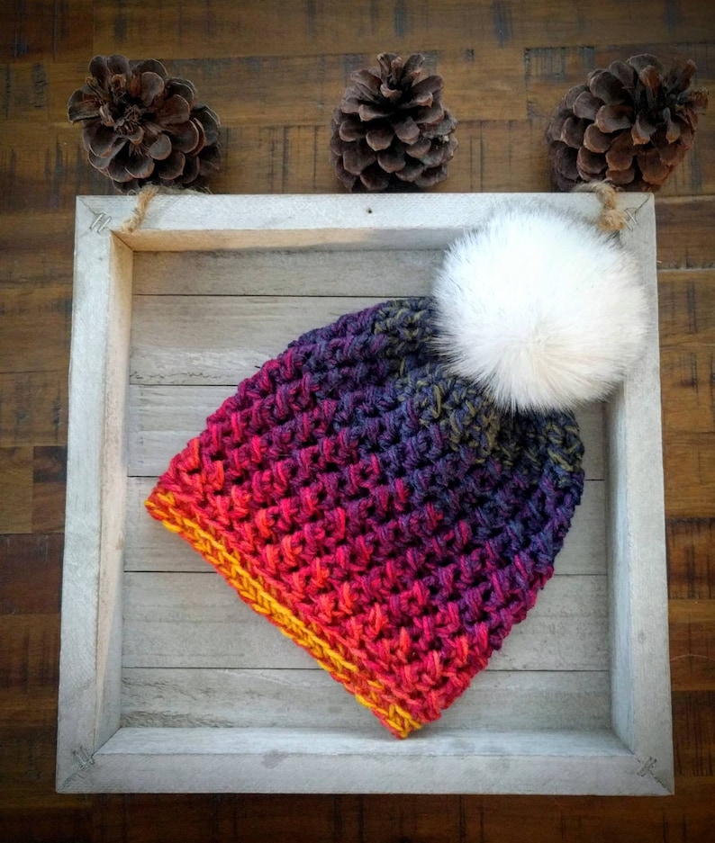 May include: A crocheted beanie with a gradient of red, orange, yellow, purple, and blue colours. The beanie has a white faux fur pom pom on top.