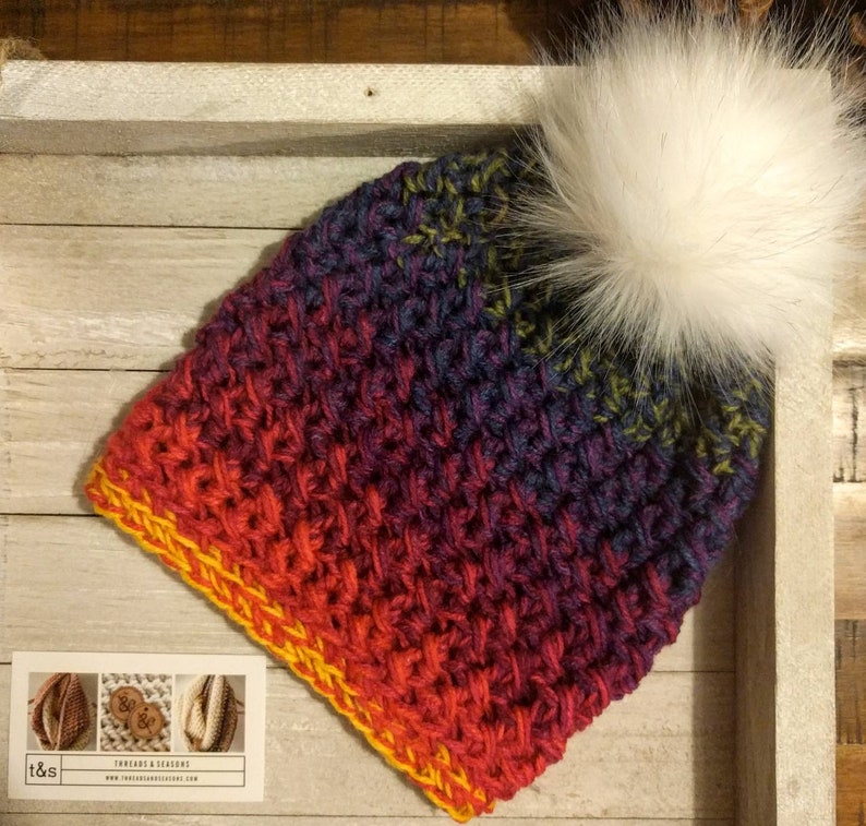 May include: A colourful crocheted beanie with a white faux fur pom pom. The beanie is made with a gradient yarn in shades of red, orange, yellow, purple, and blue. The beanie has a yellow crocheted border.