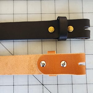 Puede incluir: Se muestran dos correas de cinturón de cuero, una negra y otra marrón claro. La correa negra tiene remaches dorados y una presilla a juego. La correa marrón claro tiene remaches plateados. Ambas correas tienen una ranura en el extremo.