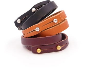 May include: Stack of leather wrap bracelets in black, tan, and burgundy. Each bracelet features a metal stud closure. The bracelets are stacked on a white background, showcasing the different colors and textures of the leather.