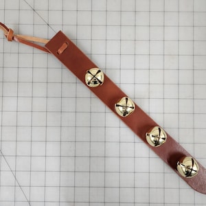 May include: A brown leather strap with four gold-colored bells attached. The strap has a loop at one end and a leather tie. The bells are evenly spaced along the strap.