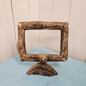 May include: A rustic, rectangular wooden picture frame with a natural, unfinished appearance. The frame is supported by a triangular base, and the wood has visible knots and grain. The frame sits on a light blue surface.
